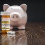 Non-Proprietary Prescription Medicine Bottle, Pills and Piggy Bank on Reflective Wooden Surface.
