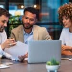 group of three business people sitting at a desk looking at a laptop and spreadsheet